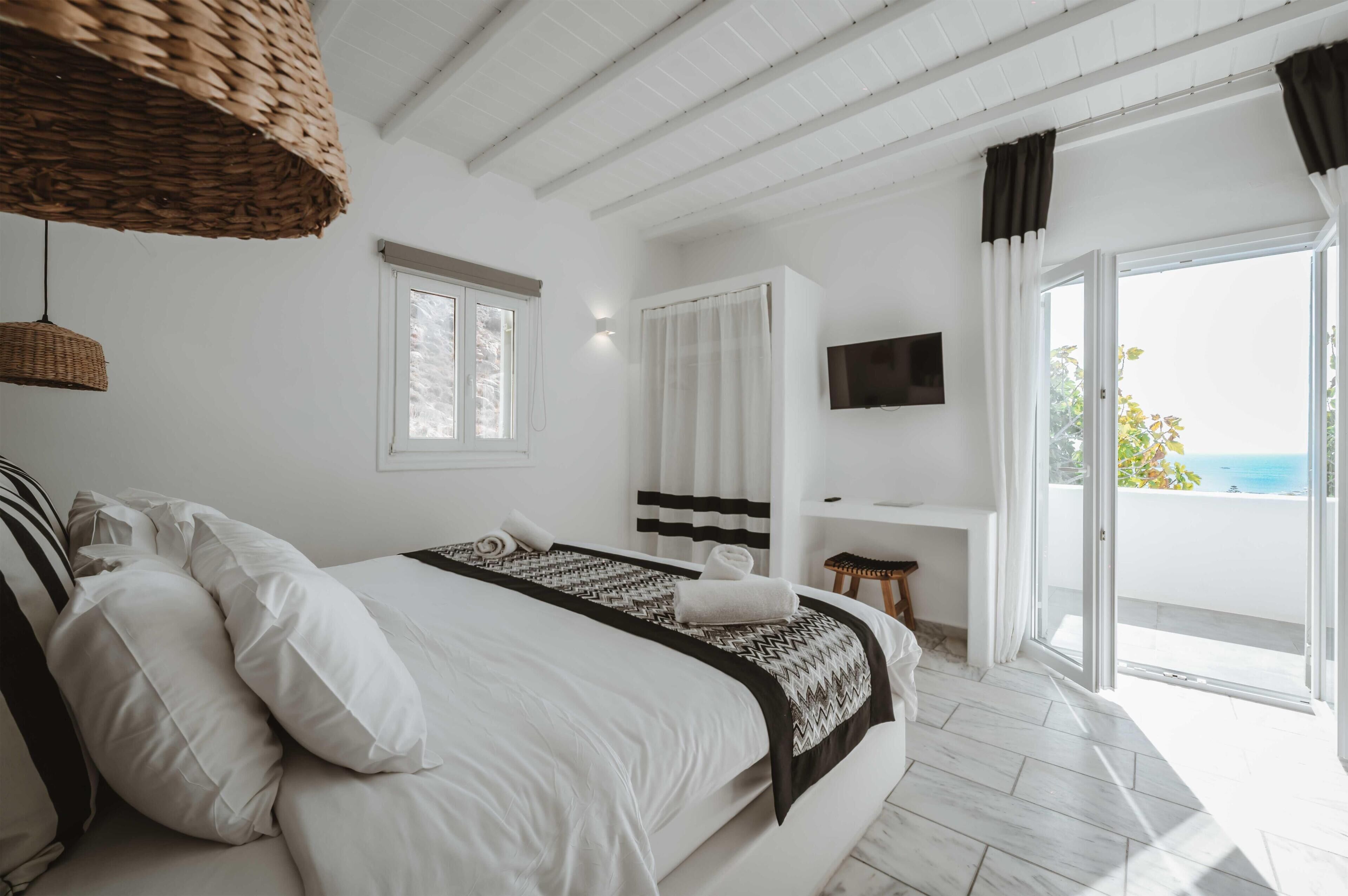 A modern bedroom with a large bed, a TV mounted on the wall, and a balcony with a view of the sea.