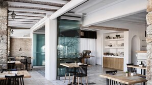 Un área de comedor moderna con mesas de madera y sillas negras, un separador de vidrio y una cocina con gabinetes y estantes blancos.