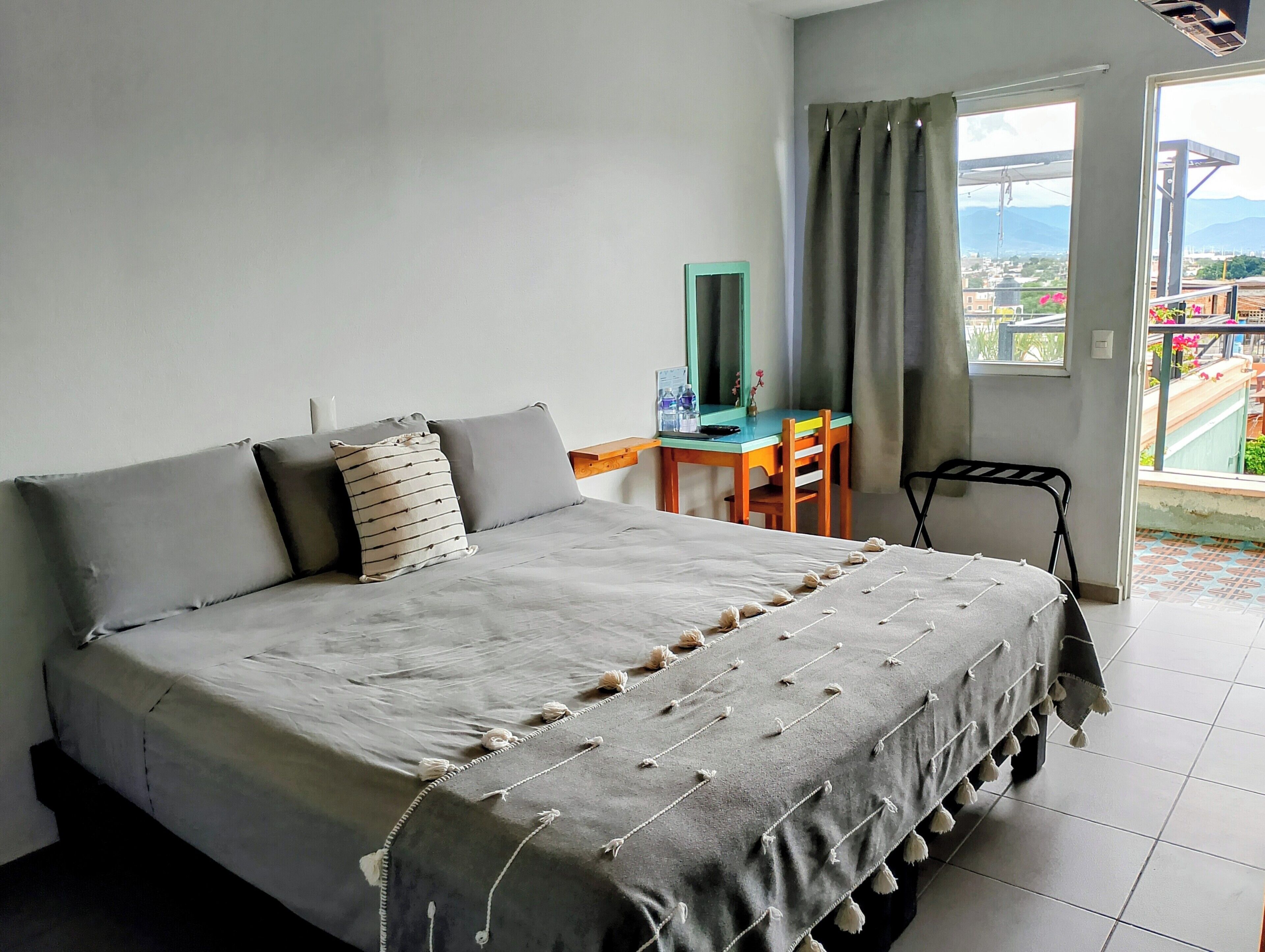 Una habitación con cama, escritorio, silla, espejo y vistas al exterior.