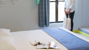 Quarto com cama, janela com cortinas, uma obra de arte na parede e um chapéu pendurado.