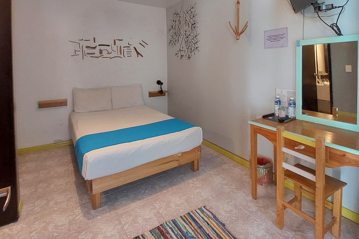Une chambre à coucher comprenant un lit, un bureau, une chaise, un téléviseur fixé au mur et un mur orné d’un élément décoratif.