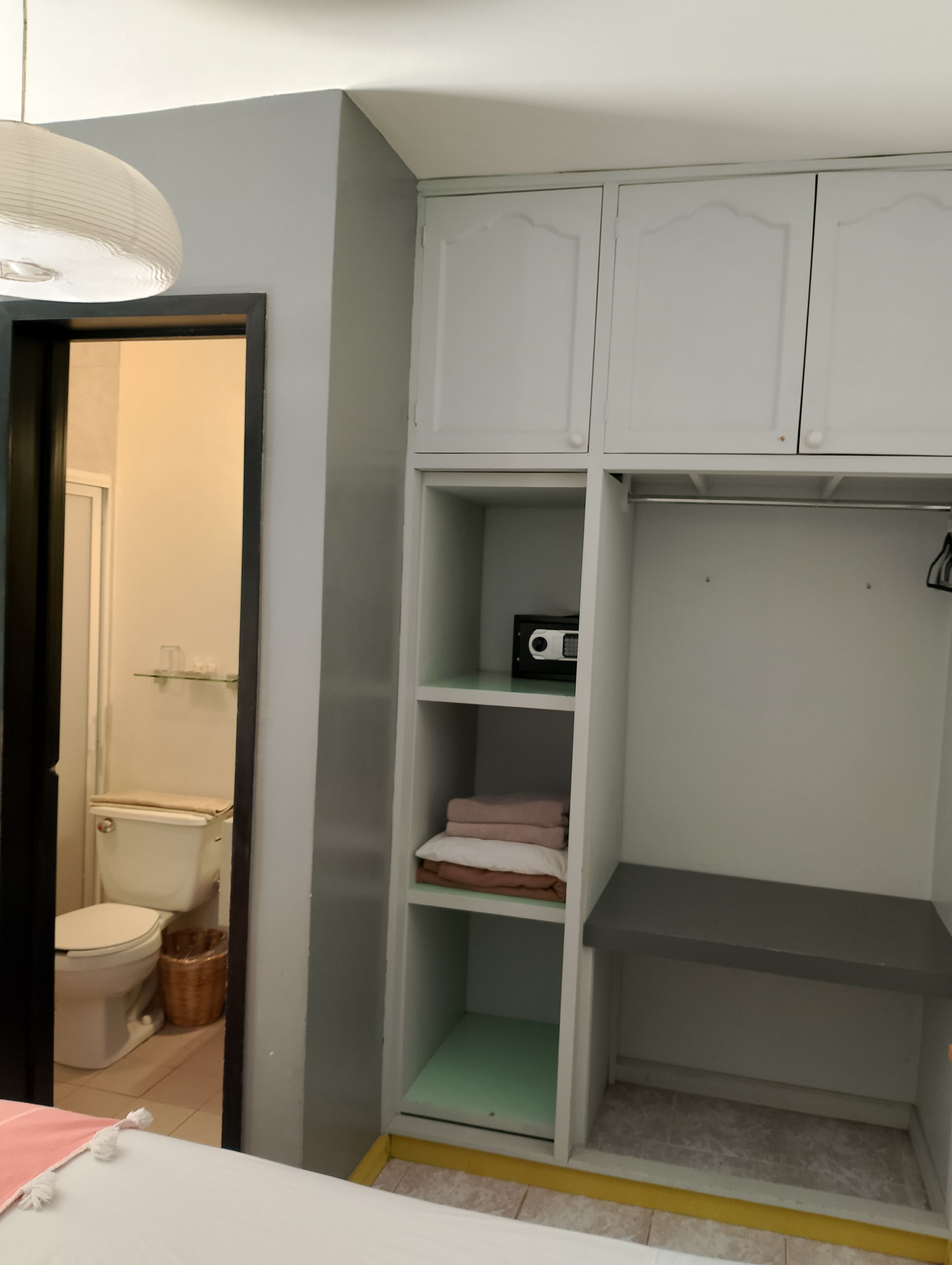 Une pièce avec une armoire blanche, un lit recouvert d’une couverture rose et une salle de bain visible à travers une porte ouverte.