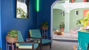 Uma área de estar ao ar livre aconchegante, com cadeiras de madeira e uma mesa, cercada por plantas em vasos e uma parede azul vibrante.