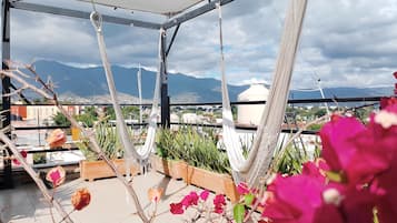 A rooftop terrace with hammocks and potted plants, overlooking a cityscape and mountains.