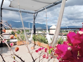 A rooftop terrace with hammocks and potted plants, overlooking a cityscape and mountains.