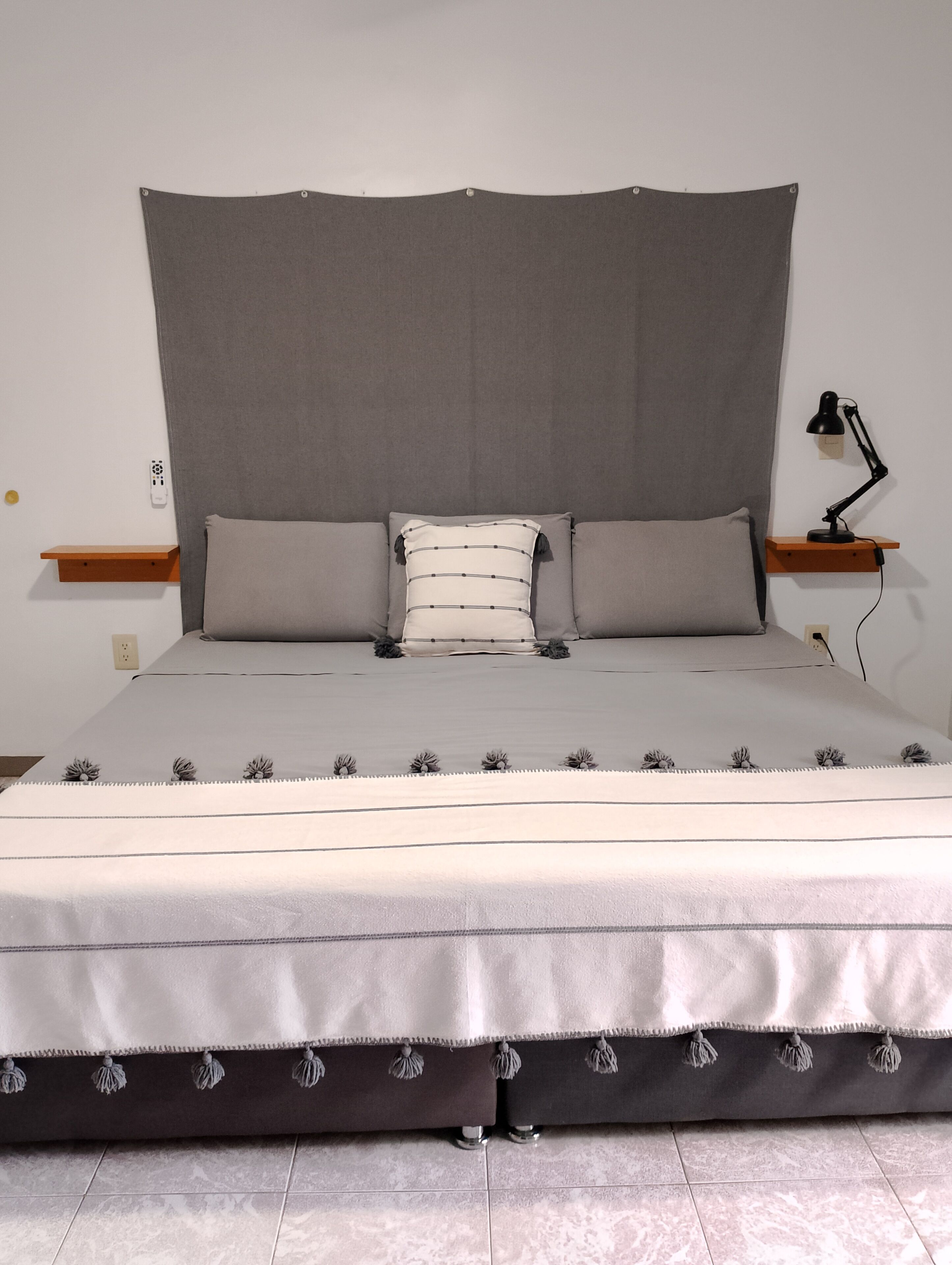 Una cama bien hecha con cabecera gris, dos almohadas, una mesita de noche con lámpara y una pequeña estantería.