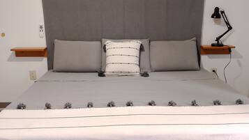 Una cama bien hecha con cabecera gris, dos almohadas, una mesita de noche con lámpara y una pequeña estantería.