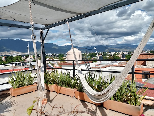 A rooftop terrace with a hammock, potted plants, and a view of a cityscape and mountains.