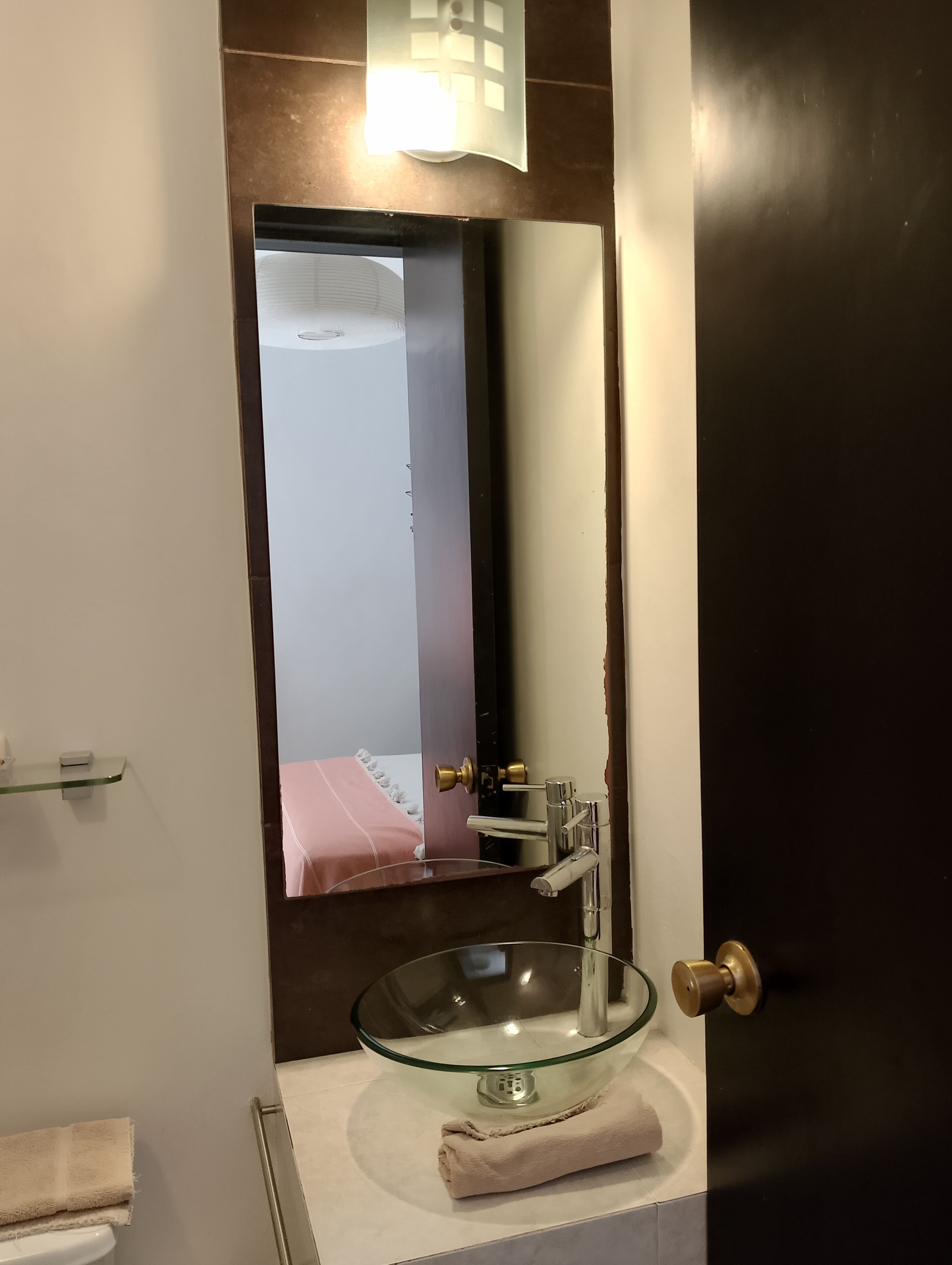 Un baño con un lavabo de vidrio, una toalla y un espejo que refleja una cama.