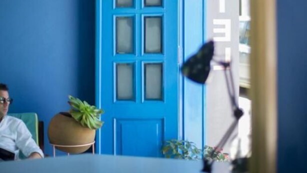 Um quarto com porta azul, uma escrivaninha, uma cadeira e um vaso de plantas.