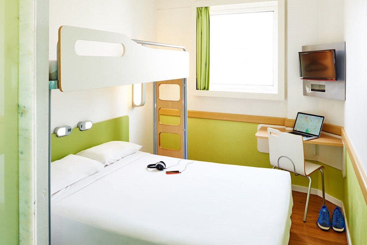 Habitación de hotel con cama, escritorio con una computadora portátil, televisor y ventana con cortinas.