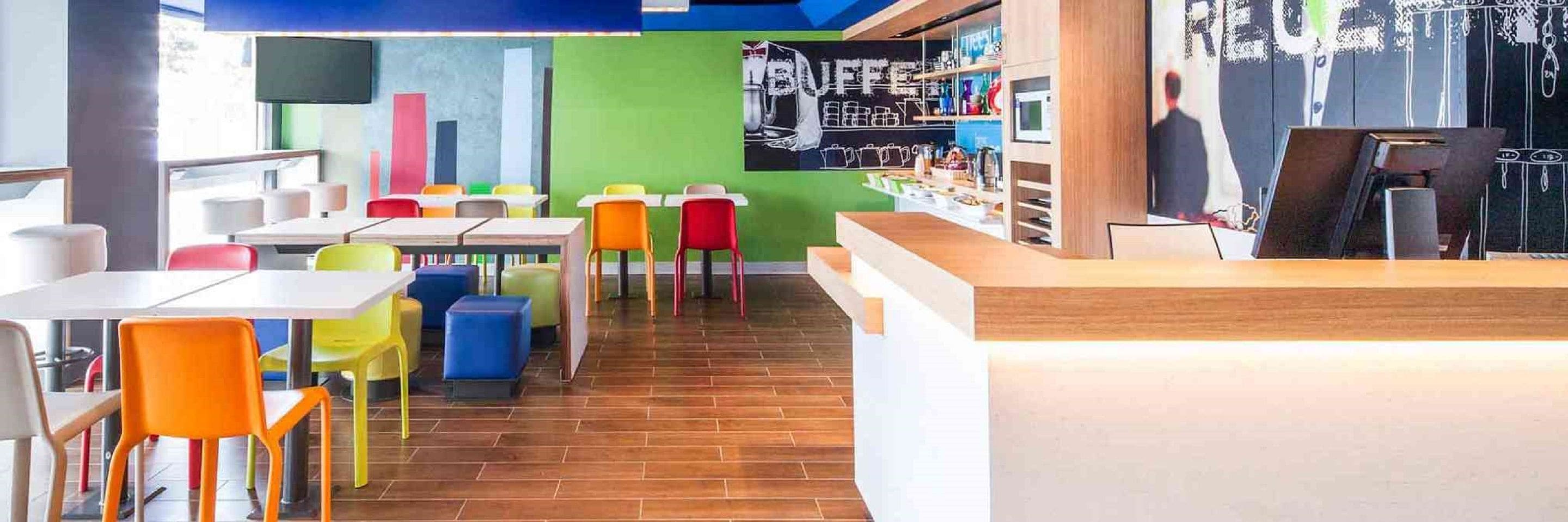 Interior moderno de un restaurante con sillas y mesas coloridas, una barra con un menú y una pared de pizarra con artículos del menú.