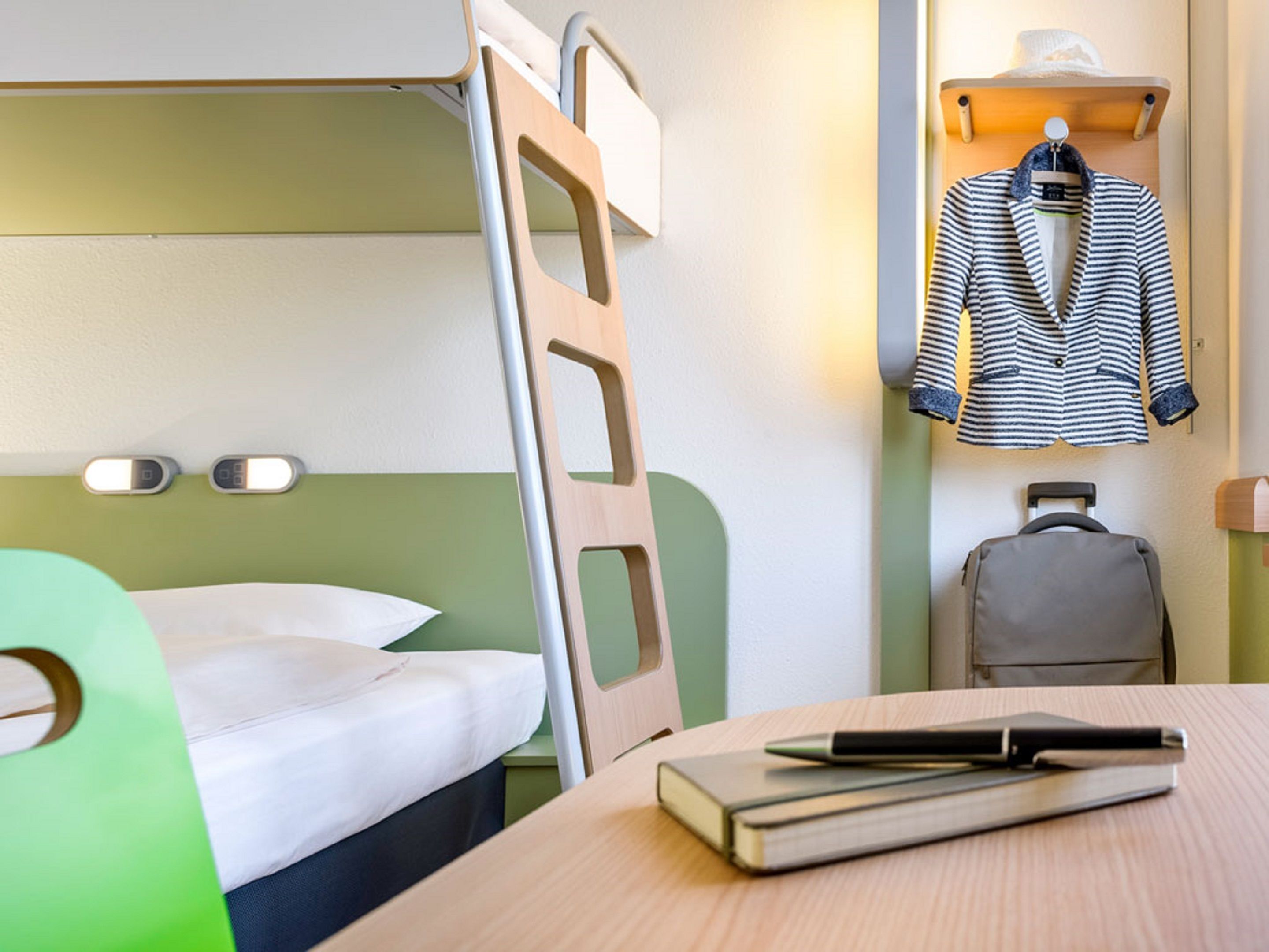 A bunk bed with a green headboard, a bedside table with a book and pen, a suitcase, and a striped jacket hanging on a hook.