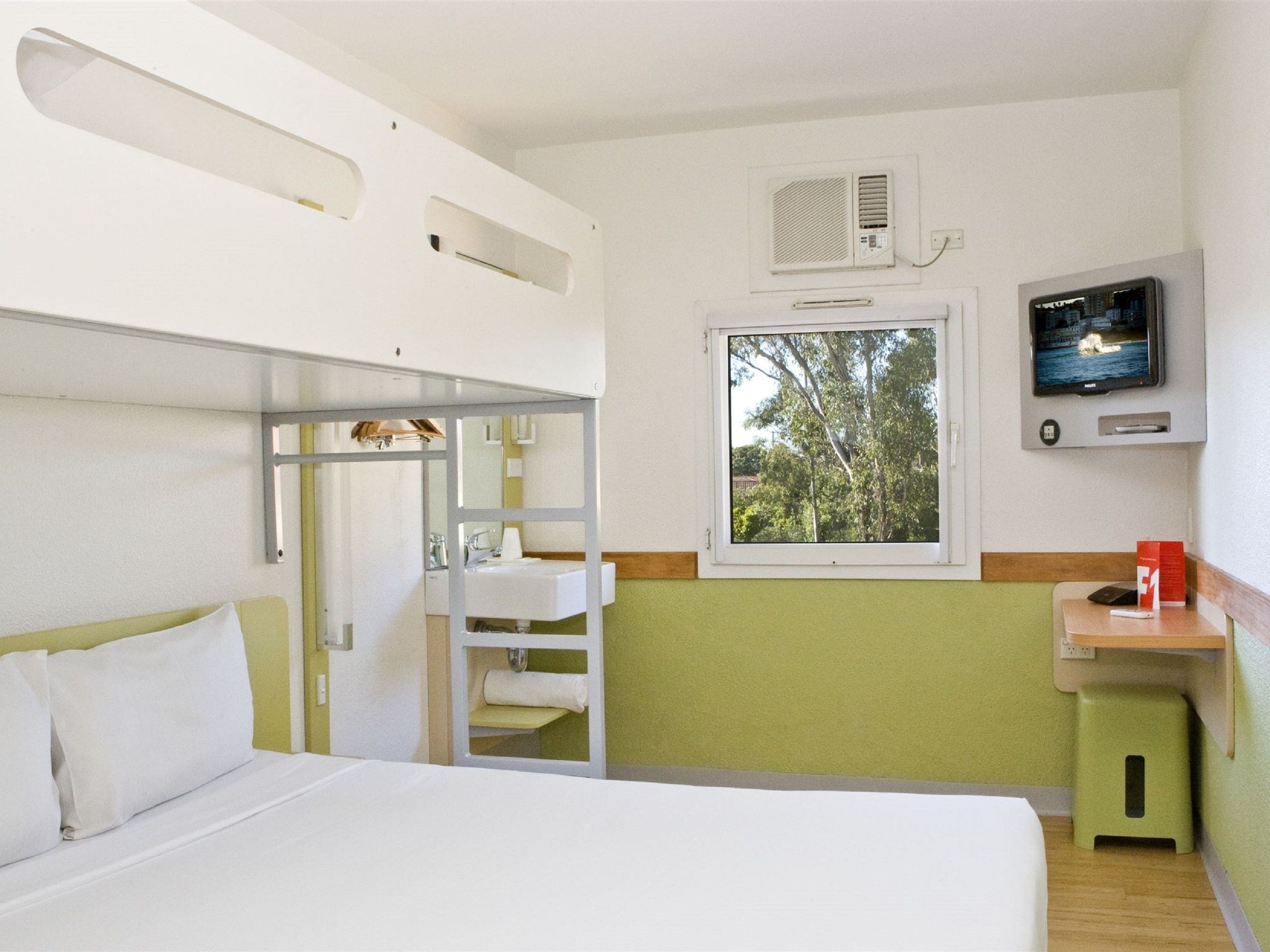 A compact hotel room with a bunk bed, a small sink, a television, and a window with a view of trees.