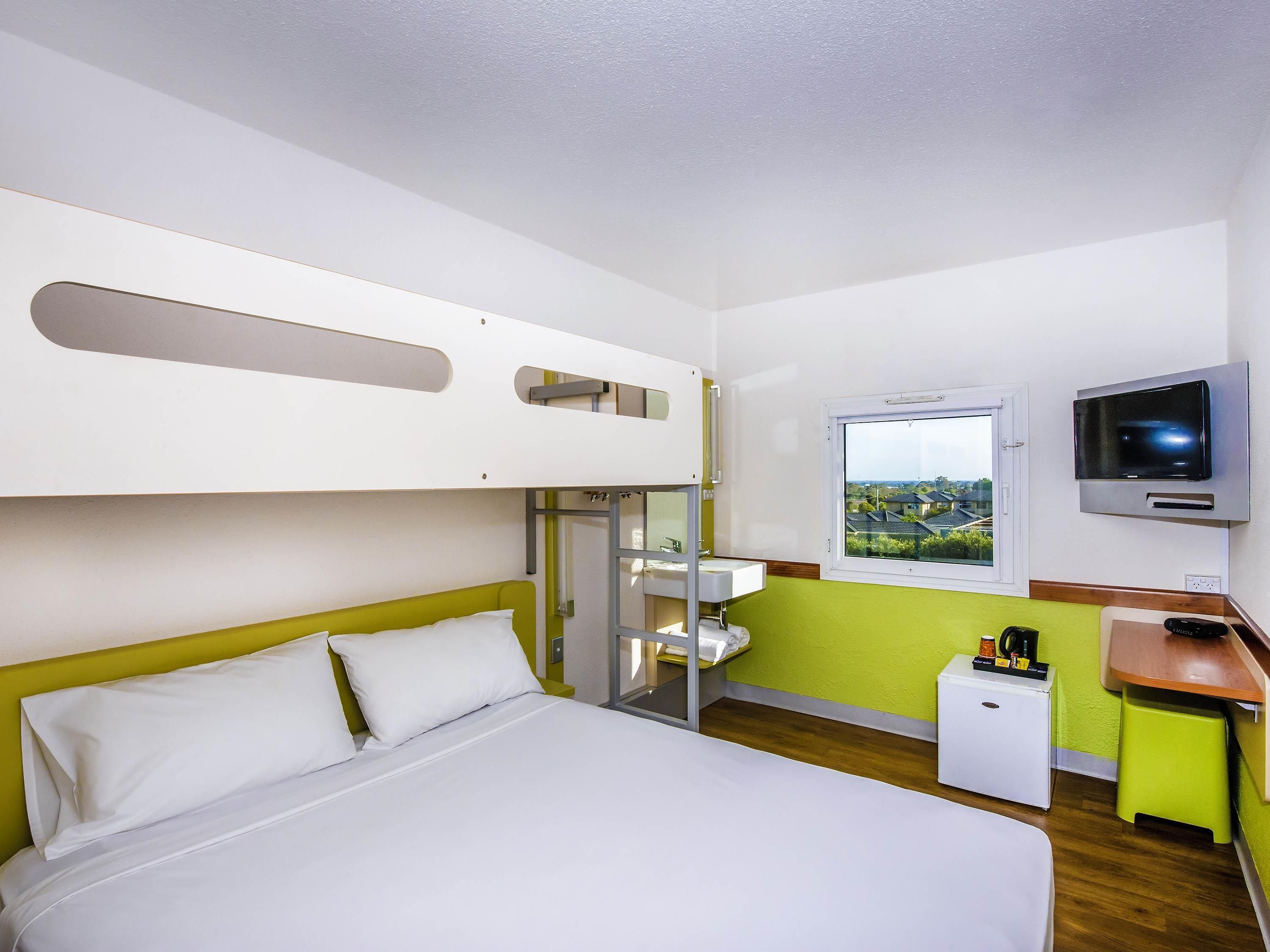 Ein Hotelzimmer mit Etagenbett, einem Einzelbett, einem Fernseher, einem Fenster mit Blick und einem kleinen Schreibtisch mit Kaffeemaschine.