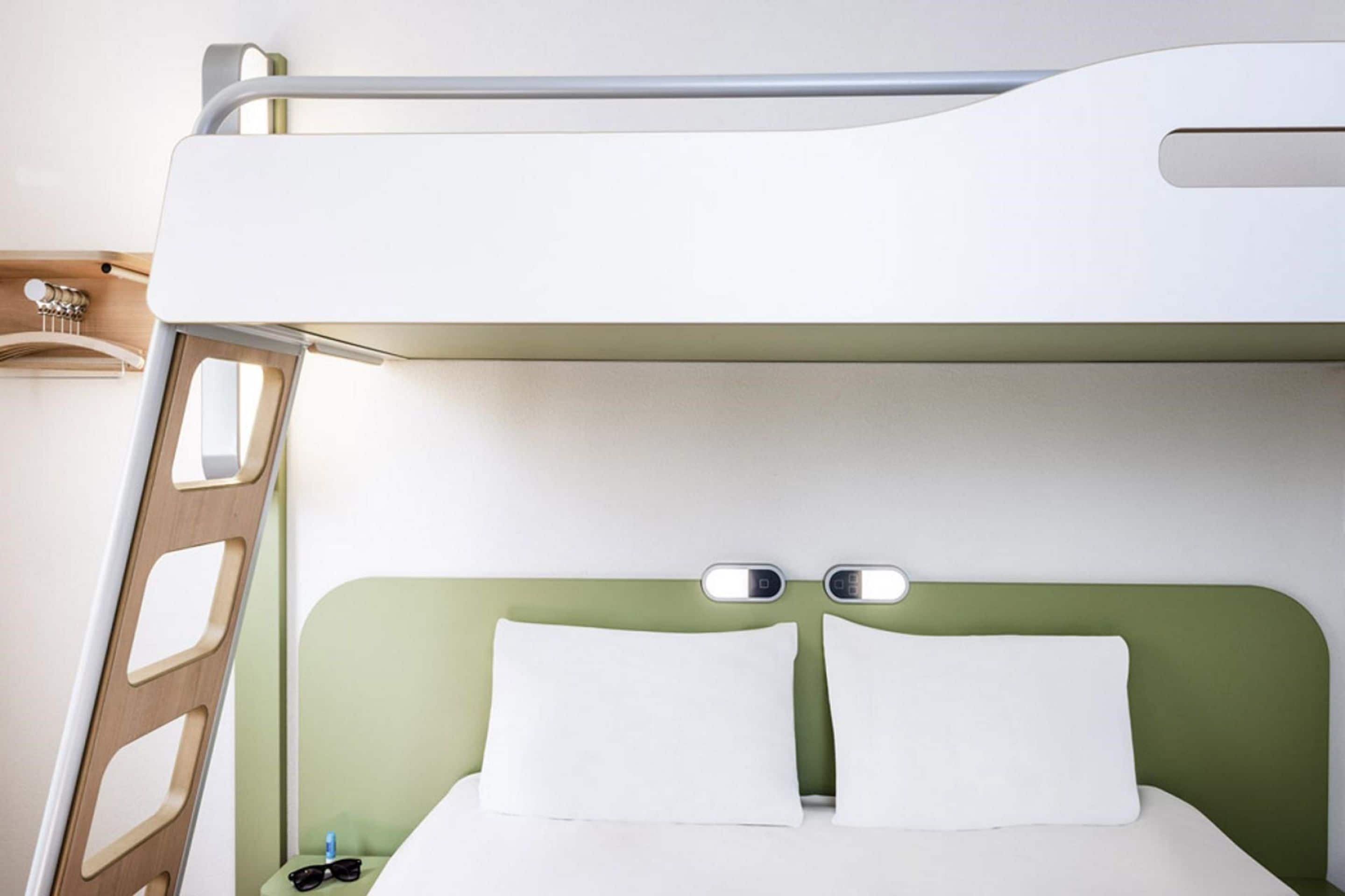 Una cama litera con cabecera verde, una escalera de madera y una mesita de noche con una botella y gafas de sol.