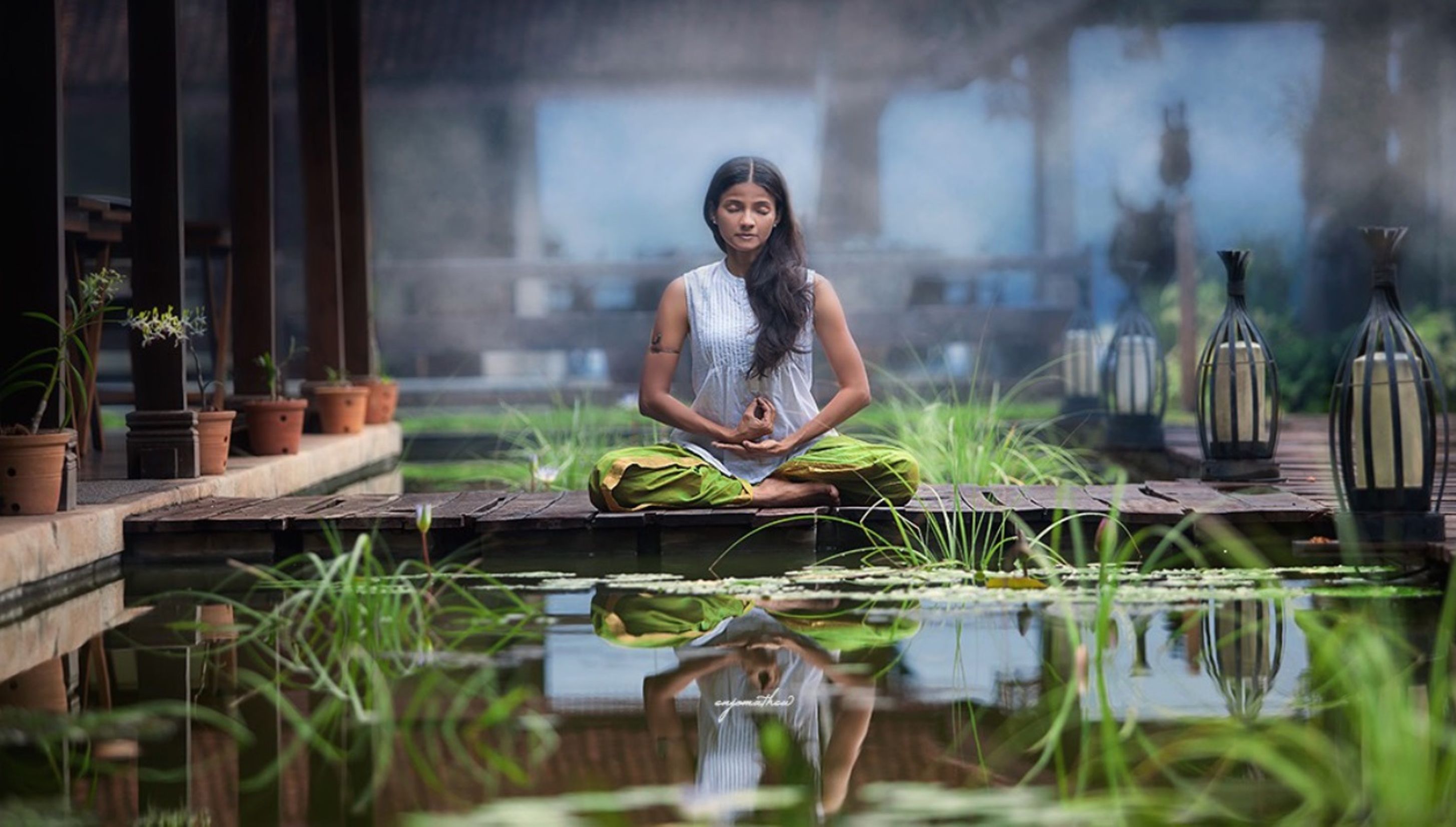 A person meditating in a serene outdoor setting with a pond, potted plants, and lanterns.