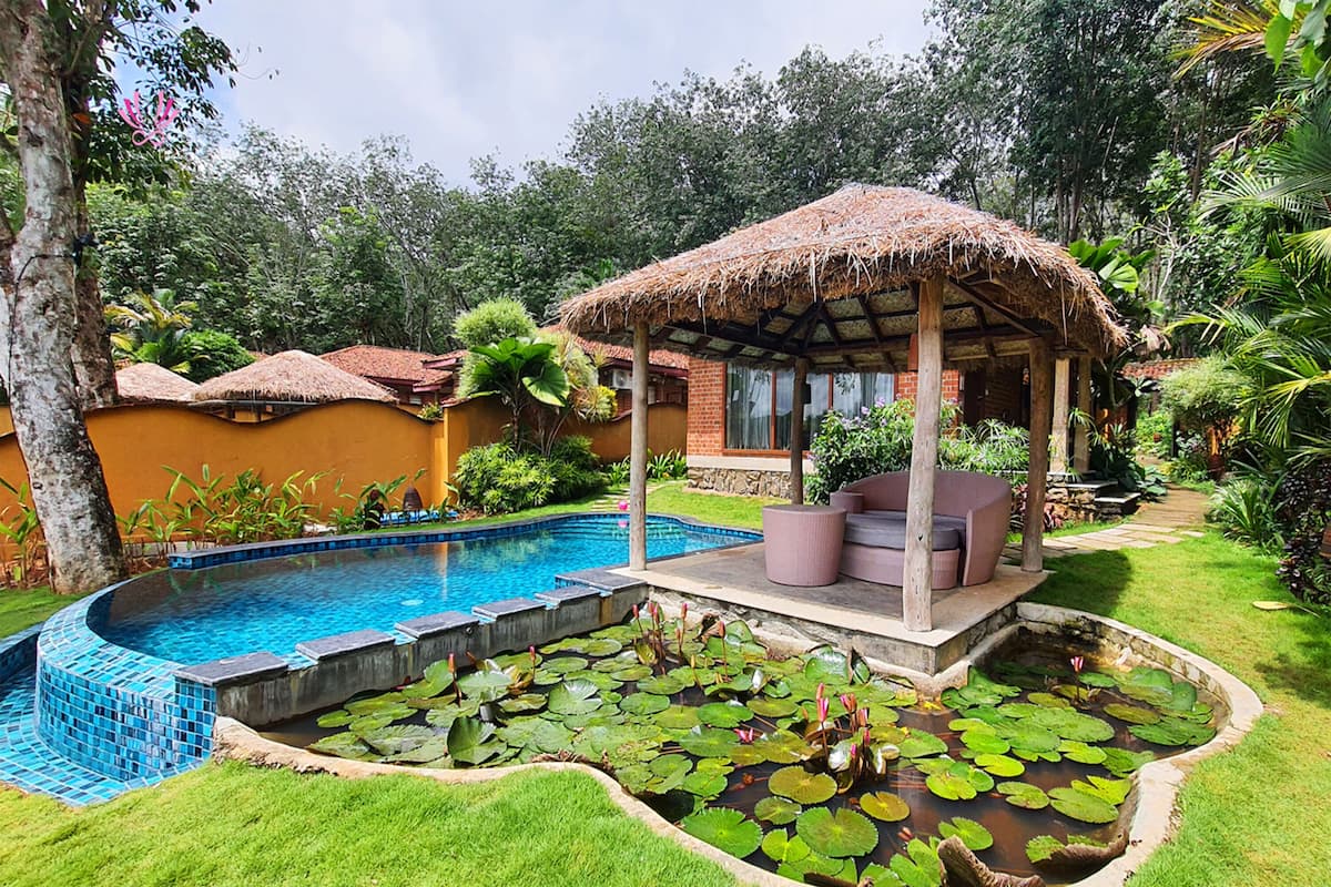 A pool with a thatched gazebo, surrounded by lush greenery and a small garden area with lily pads.