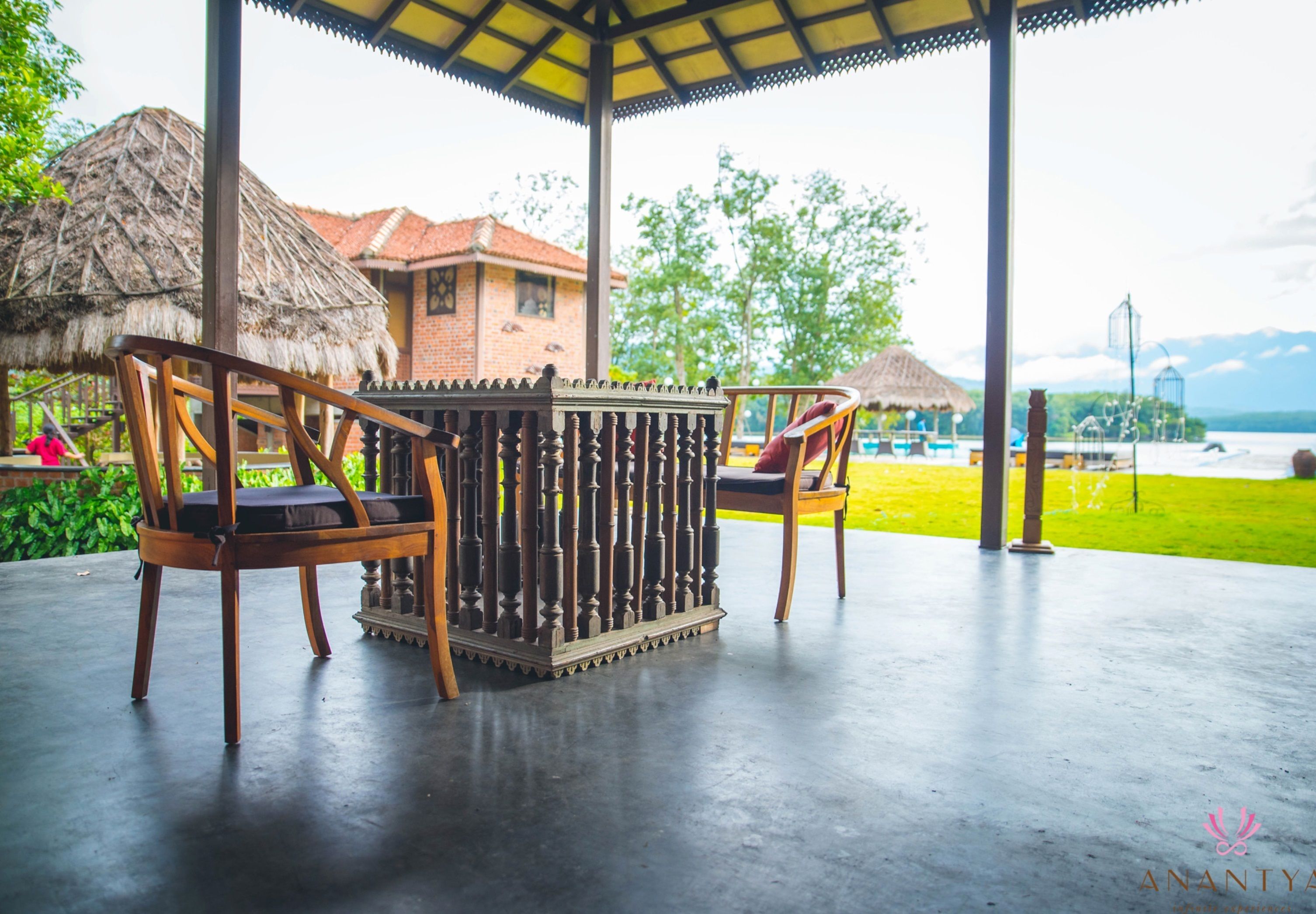 A gazebo with two wooden chairs and a table, overlooking a grassy area and a building with a red roof.