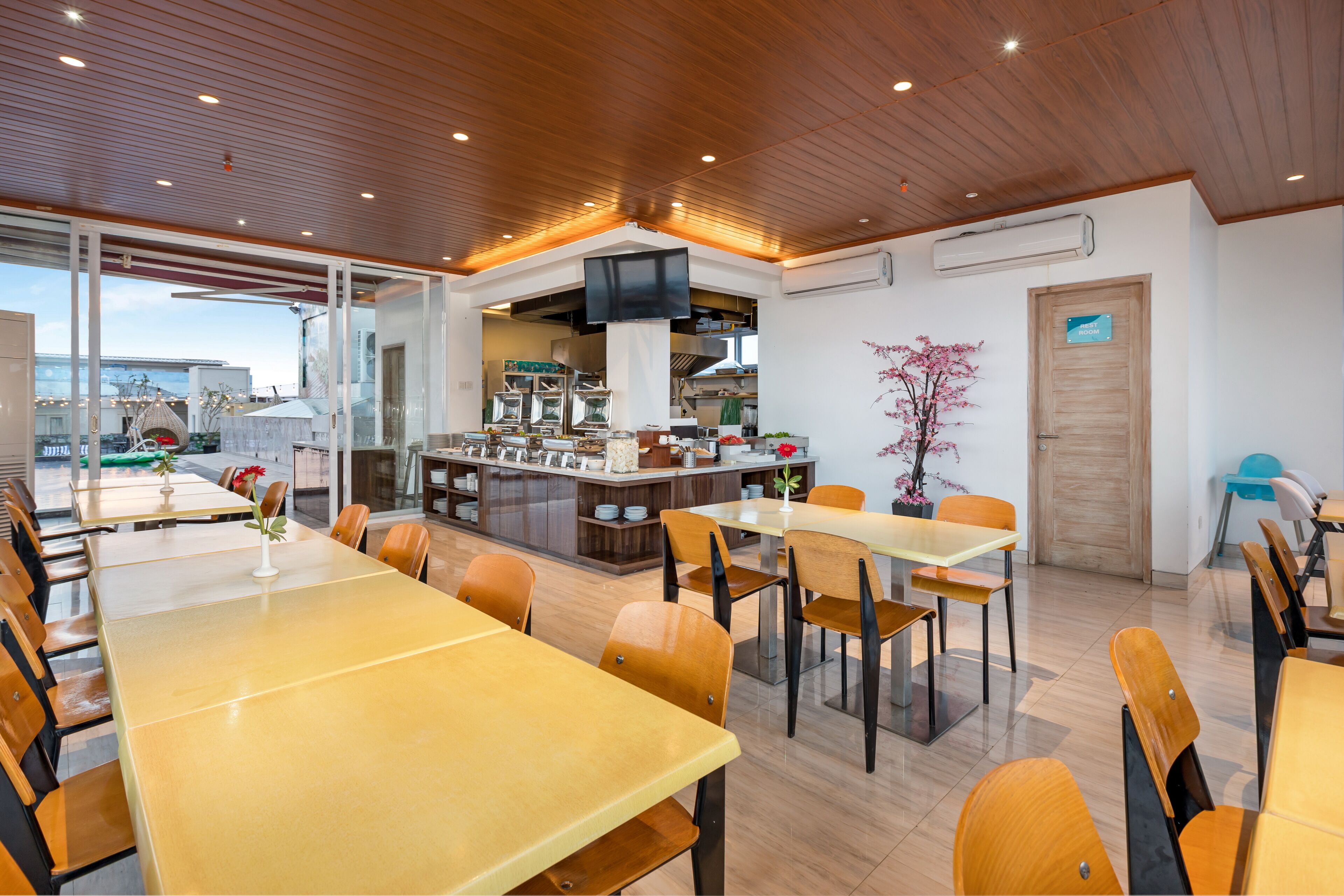 Un comedor moderno con mesas y sillas de madera, una cocina al fondo y vistas al exterior.