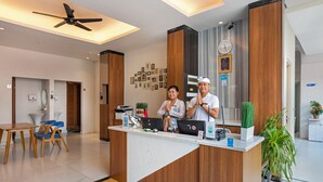 A reception desk with a modern design, featuring a wooden panel and a white counter. There are two people behind the desk, one appears to be a receptionist. The room has a ceiling fan, a clock, and framed pictures on the wall. - Boss Legian Hotel Powered by Archipelago (Legian)