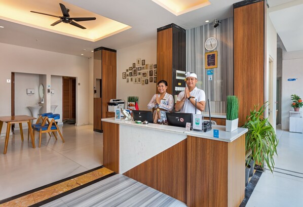 A reception desk with a modern design, featuring a wooden panel and a white counter. There are two people behind the desk, one appears to be a receptionist. The room has a ceiling fan, a clock, and framed pictures on the wall. - Boss Legian Hotel Powered by Archipelago (Legian)