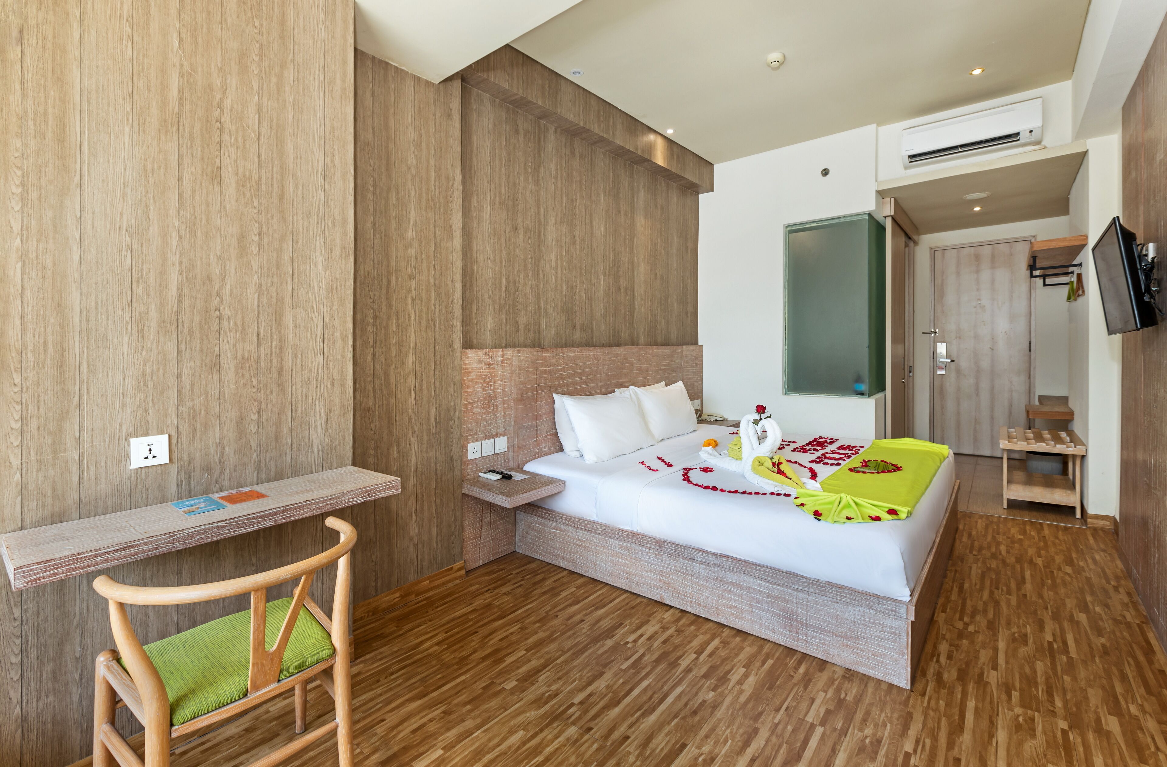 A modern hotel room with a wooden floor, a bed with white linens, a wooden desk, and a TV mounted on the wall.