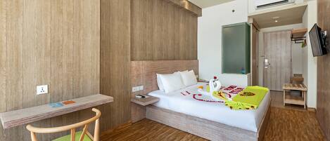 Una habitación de hotel moderna con piso de madera, una cama con ropa blanca, un escritorio de madera y un televisor montado en la pared.