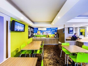 Un café moderno con sillas y mesas de color verde brillante, una barra con diferentes productos alimenticios y una pantalla de televisión mostrando una foto grupal.