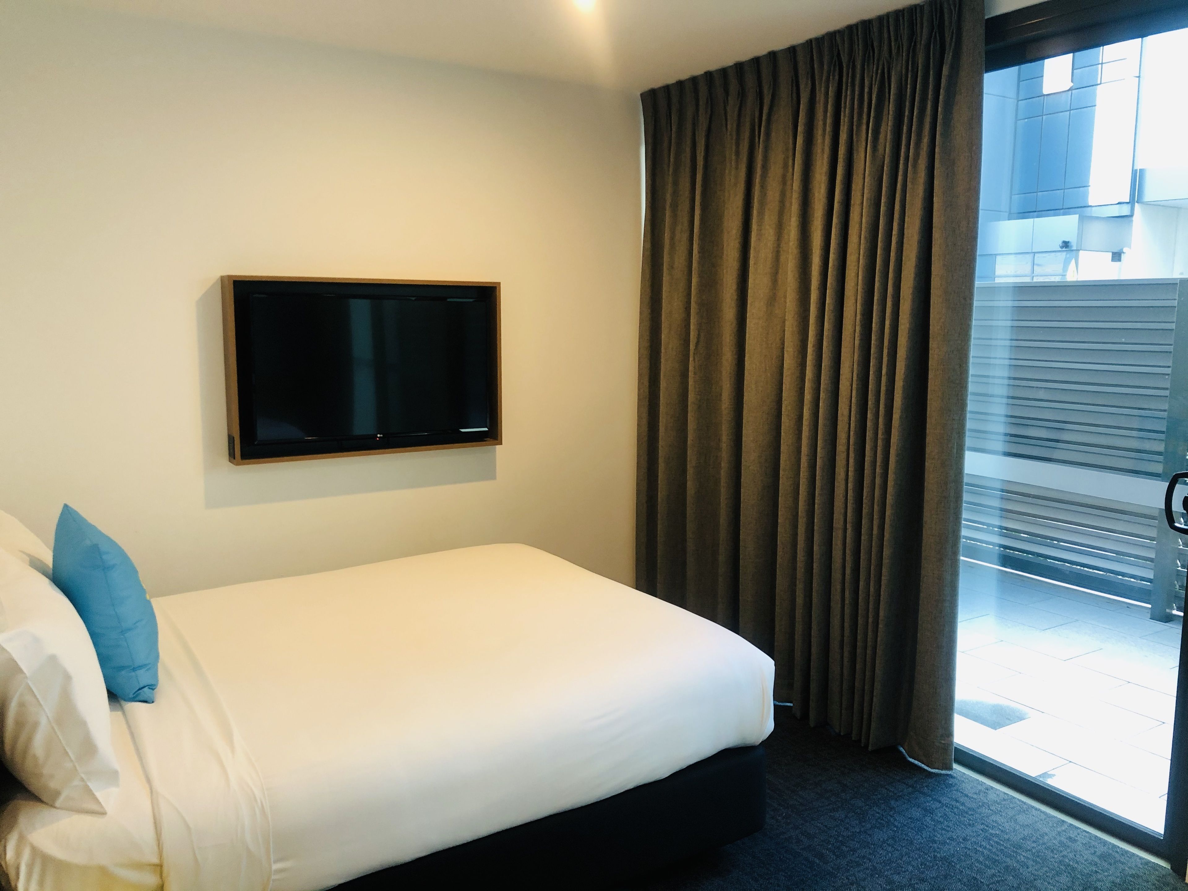 A hotel room with a bed, a television mounted on the wall, and a view of the outside through a window with curtains.