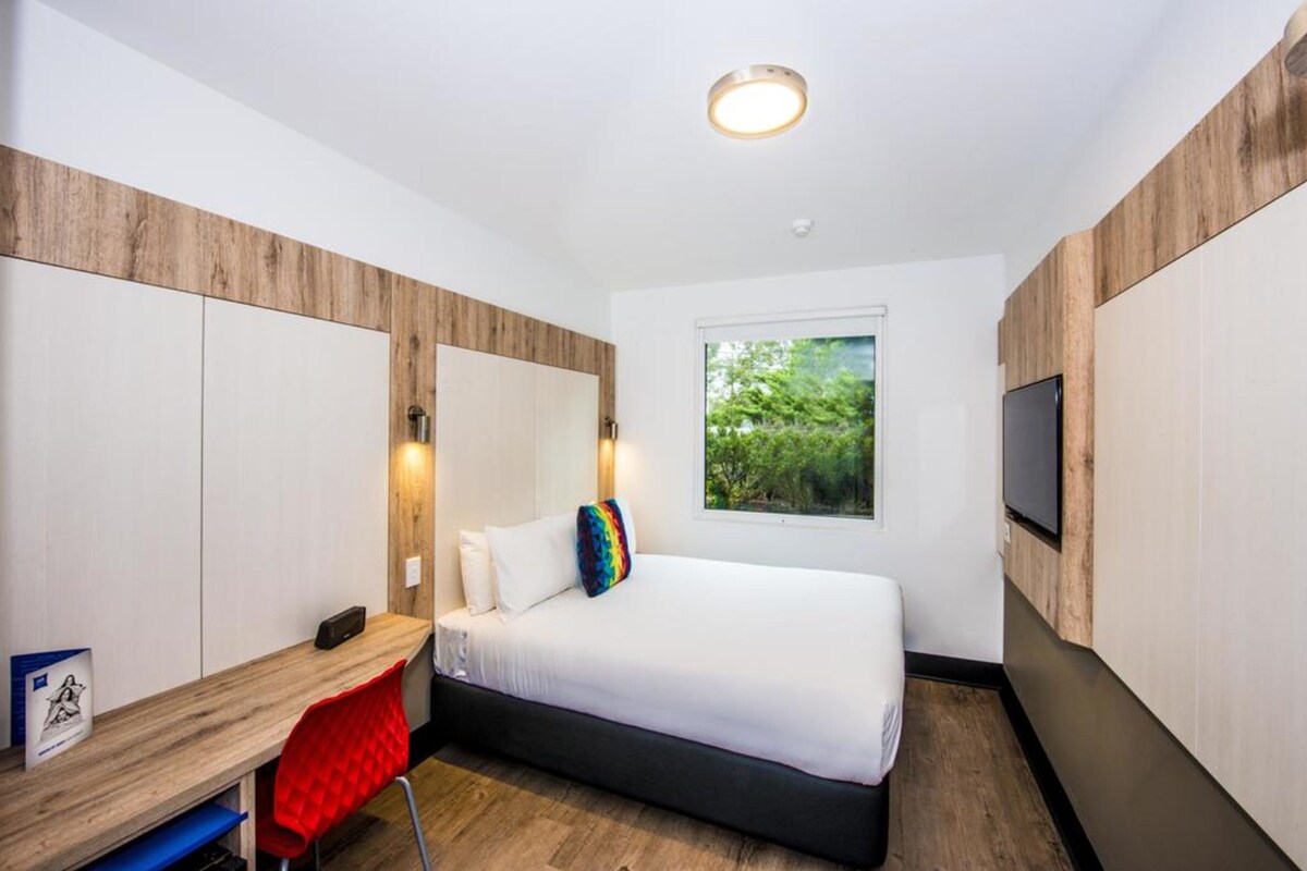 Een moderne hotelkamer met een bed, een bureau, een stoel, een televisie en een raam met uitzicht op groen.