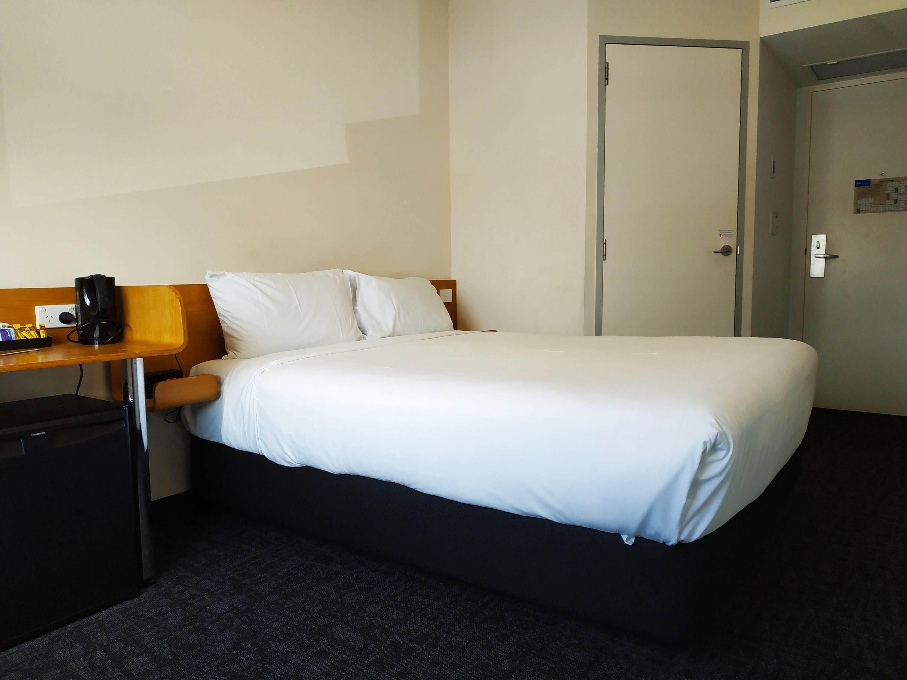 A hotel room with a bed, a desk with a chair, a small trash bin, and a door.