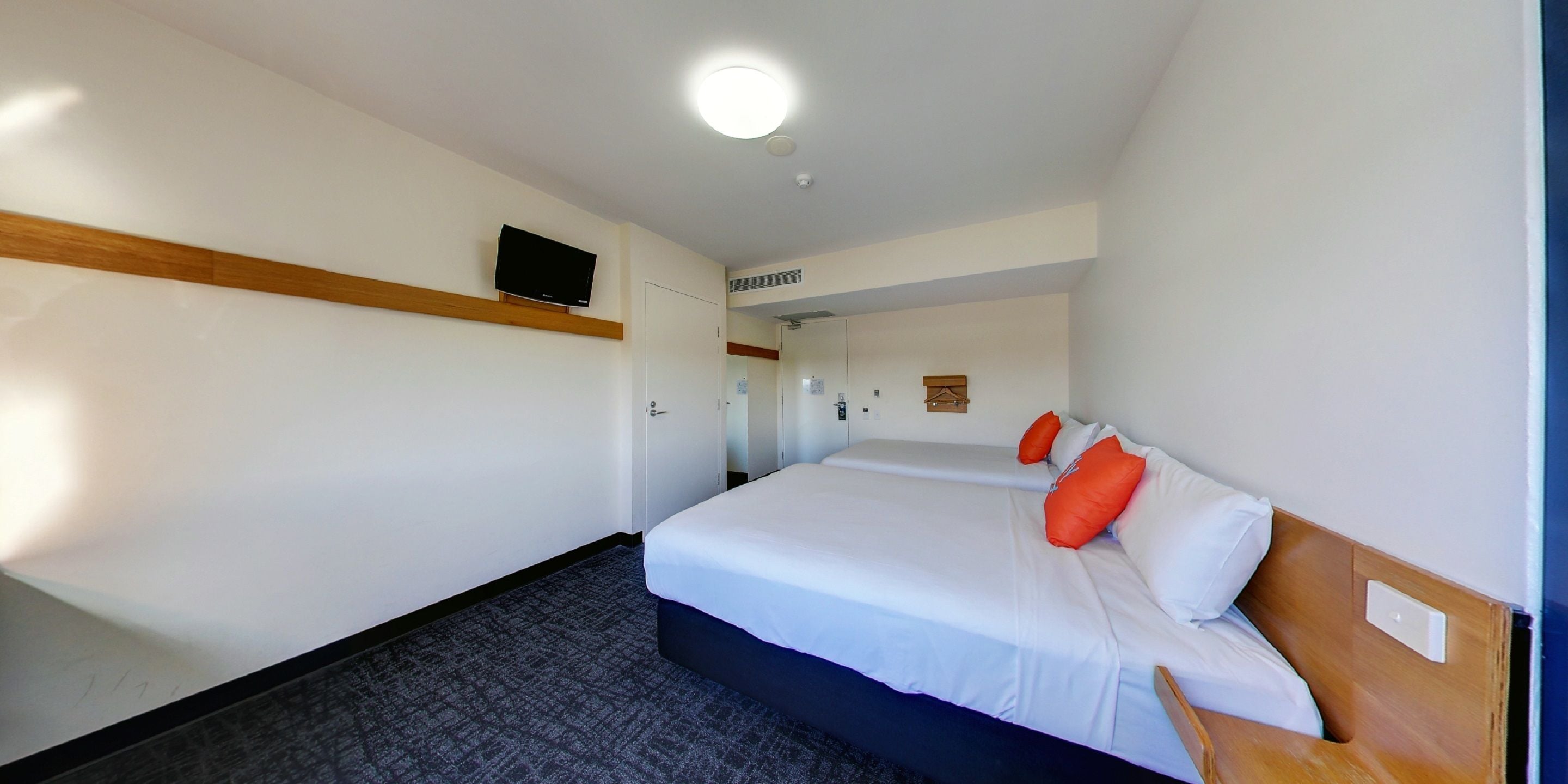 Habitación de hotel con una cama individual, un televisor de pantalla plana montado en la pared, cabecero de madera y piso alfombrado.