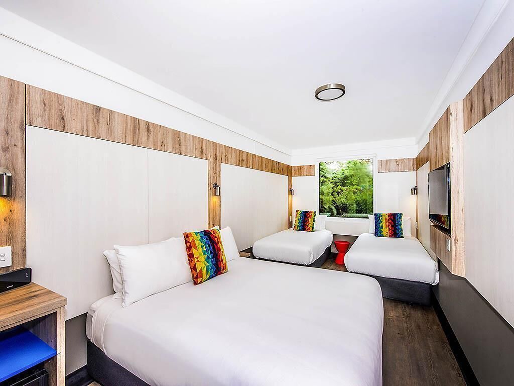 A hotel room with two beds, a TV, a desk, and a window with a view of greenery.