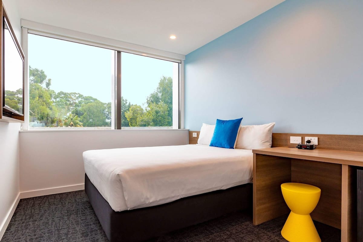 Een hotelkamer met een bed, bureau, stoel en raam met uitzicht op bomen.