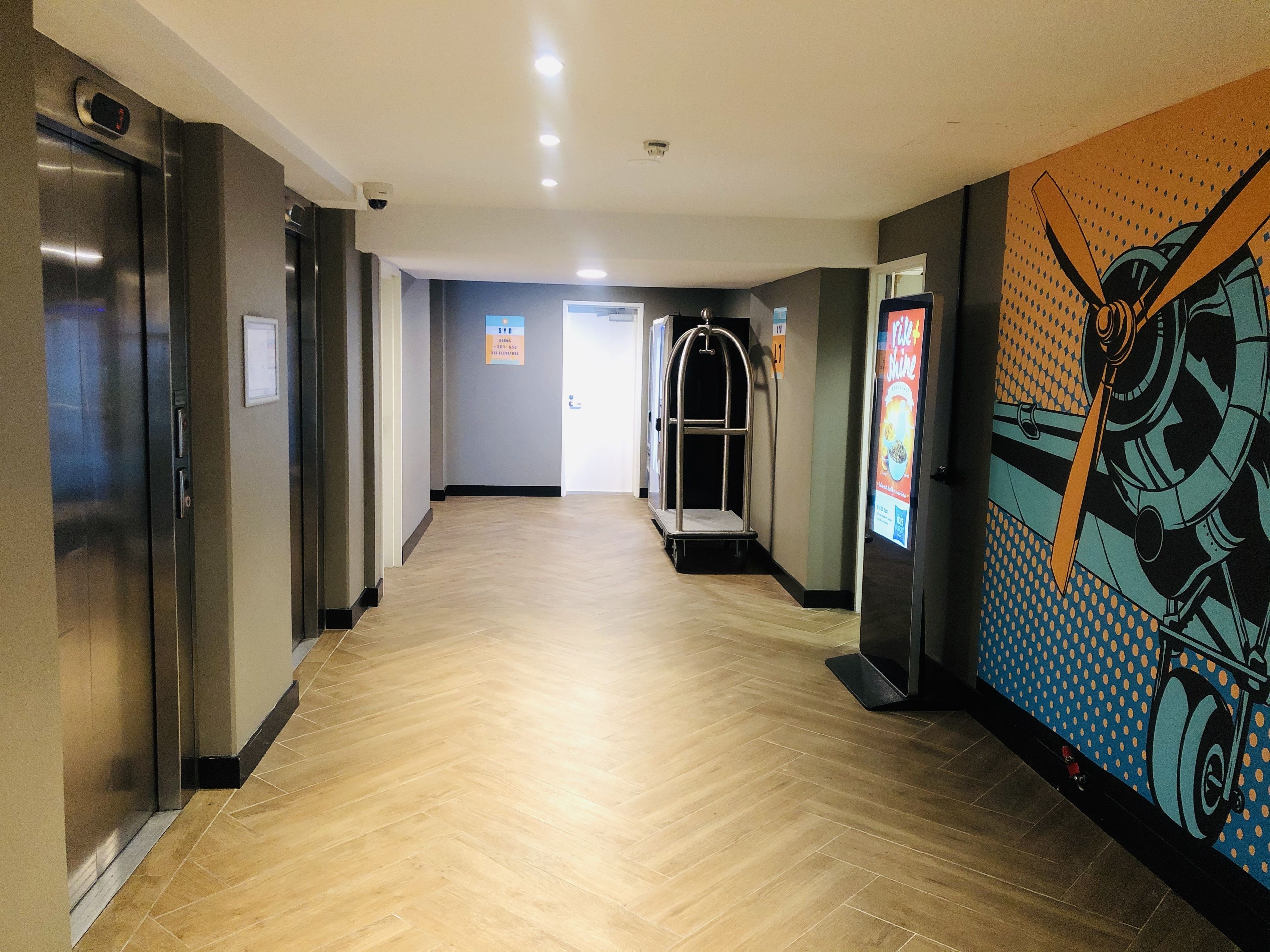 Un pasillo moderno de hotel con ascensores, un carrito de equipaje e ilustración grande y colorida en la pared.