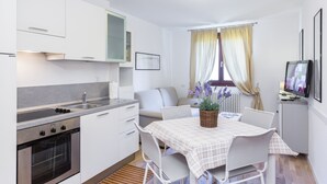 Une cuisine compacte avec des meubles blancs, un four encastré et un coin repas agrémenté d’une nappe à carreaux.