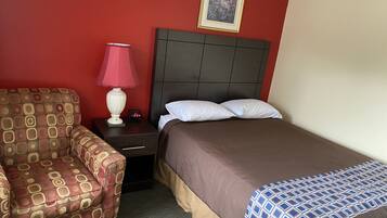 A hotel room with a patterned armchair, a bedside table with a lamp and a digital clock, a bed with a patterned bedspread, and a framed picture on the wall.