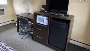 A hotel room with a desk, a television, a microwave, a refrigerator, a chair, and a lamp.