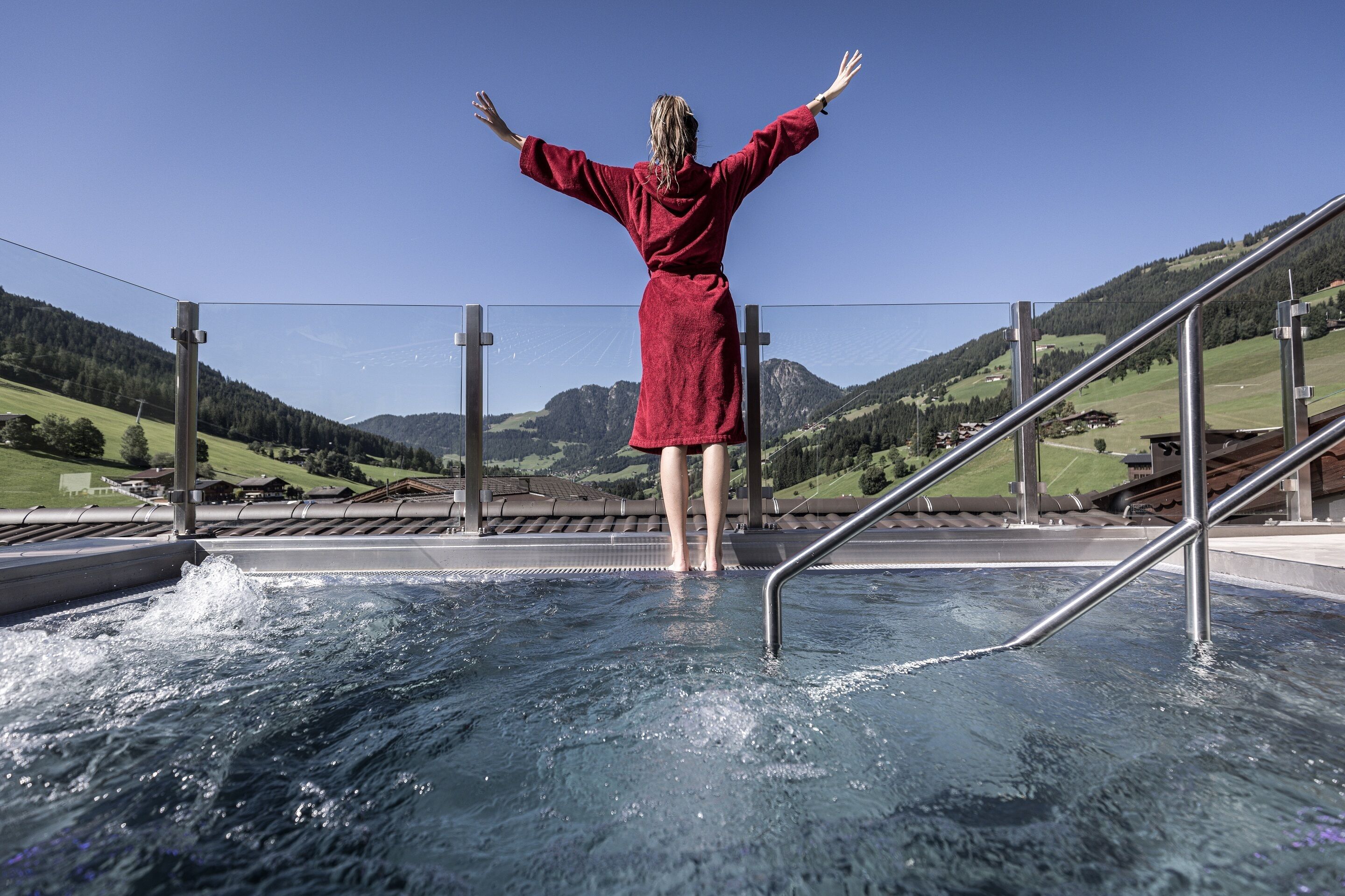 Una persona con bata roja de pie en una piscina, con los brazos extendidos, contemplando un paisaje montañoso.