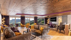 A spacious living area with a wooden ceiling, large windows offering a mountain view, and modern furniture. - Galtenberg Family & Wellness Resort (Alpbach)