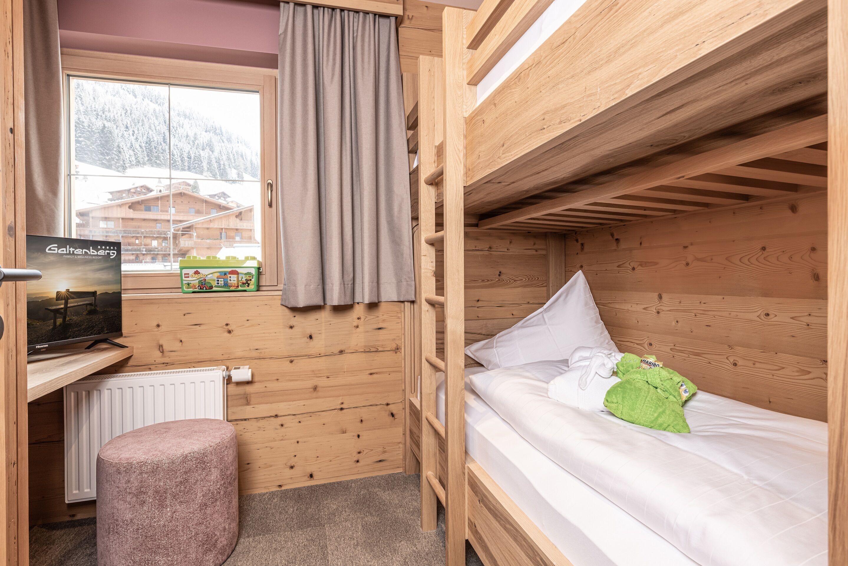 A wooden cabin room with a bunk bed, a small desk with a computer, a window with a view of a mountain, and a radiator.