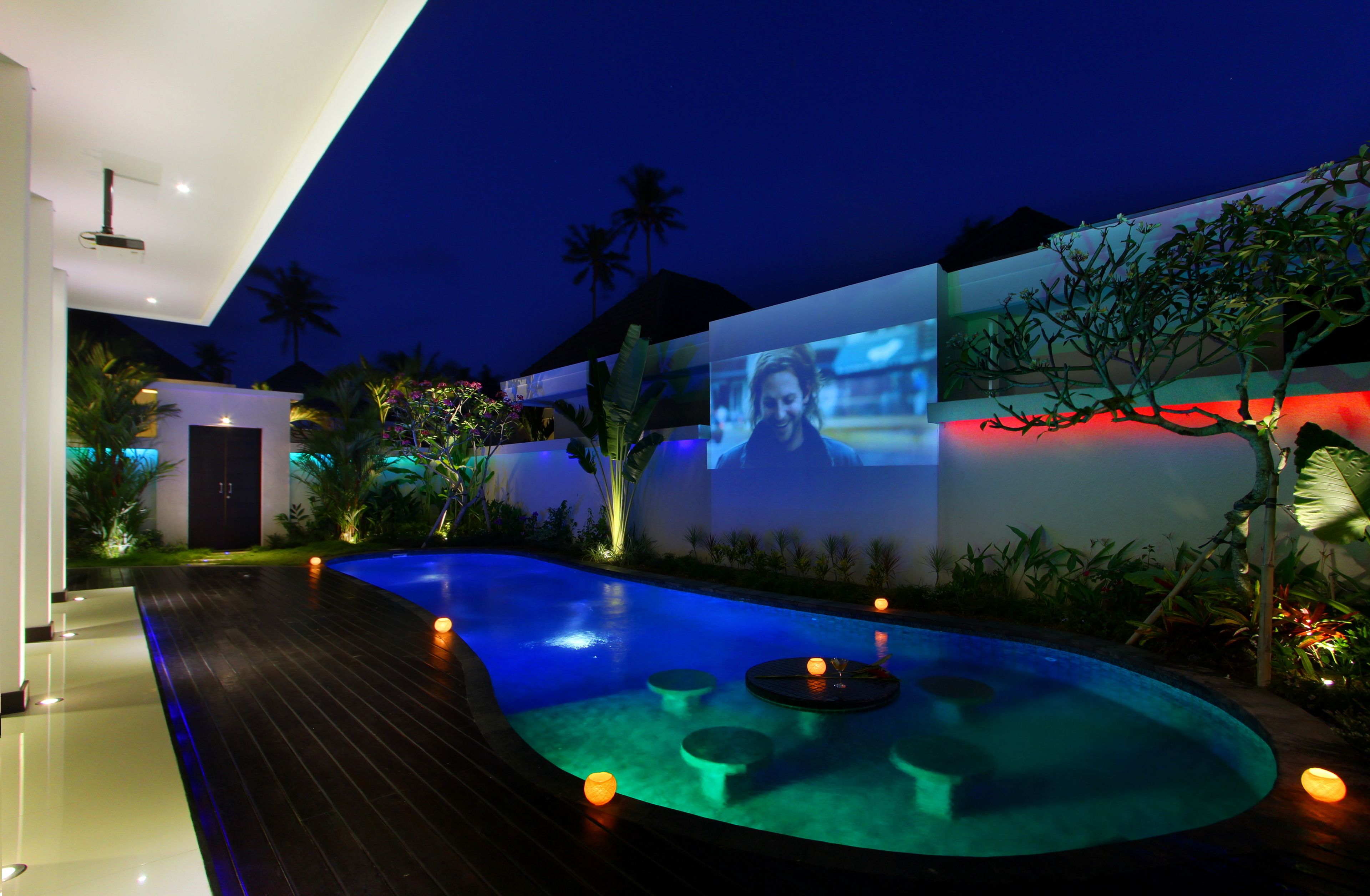 A modern outdoor pool area with a curved design, ambient lighting, and a projection screen displaying a person.