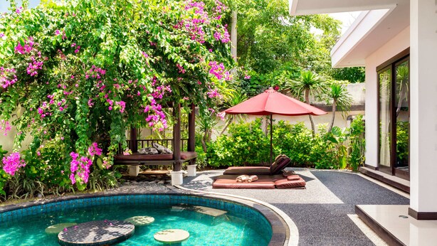 A swimming pool with a tiled edge, surrounded by a garden with pink flowers and a red umbrella. - D&G Villas (Nusa Dua)