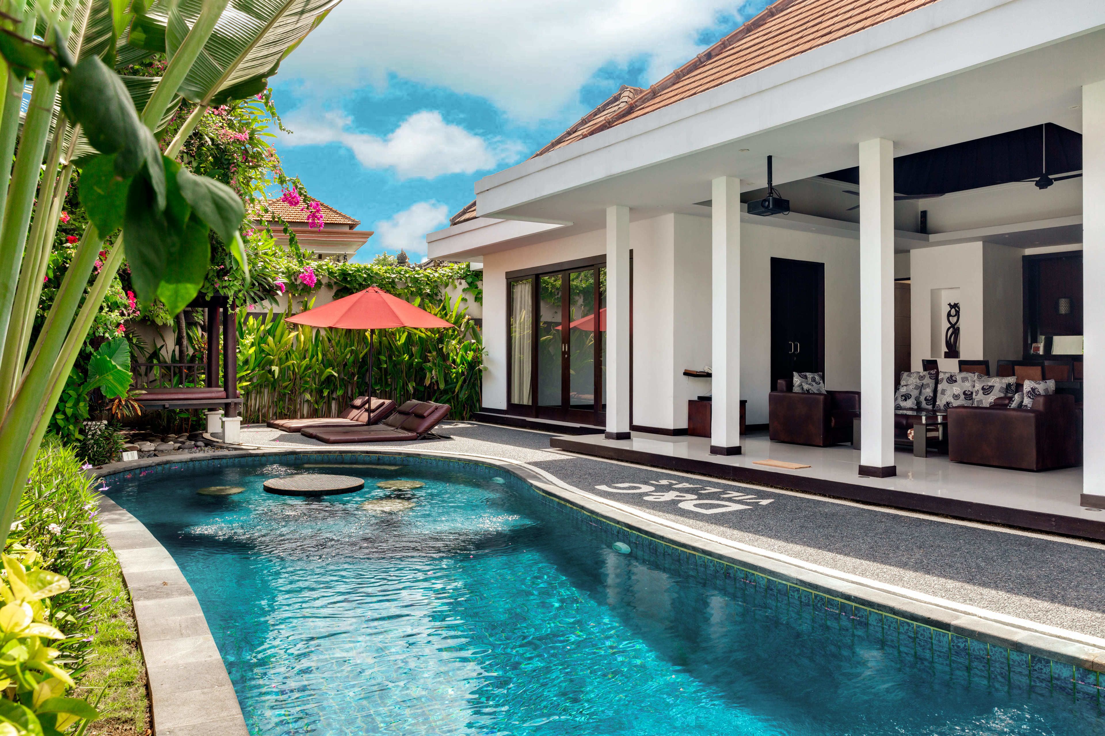 A swimming pool with a lounge area, a covered patio, and a view of a house with a tiled roof.