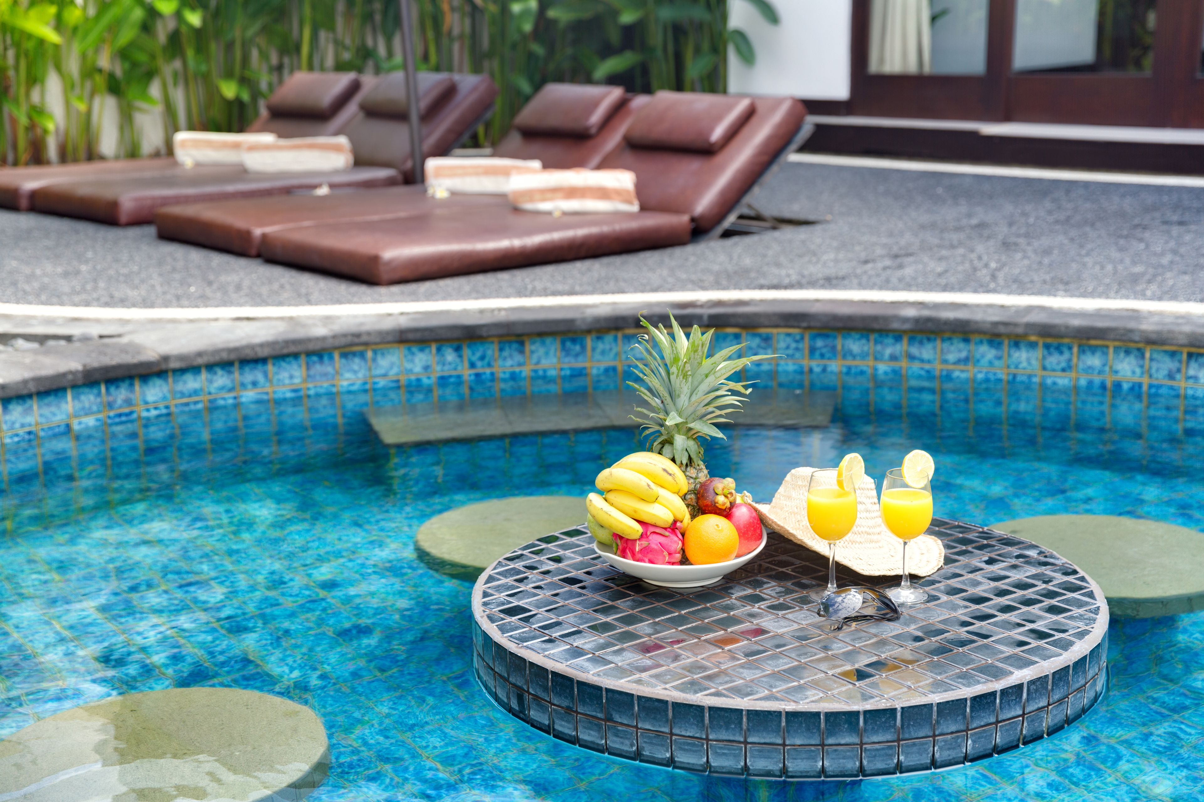 A poolside area with lounge chairs, a tiled table with fruit and drinks, and a clear blue pool.