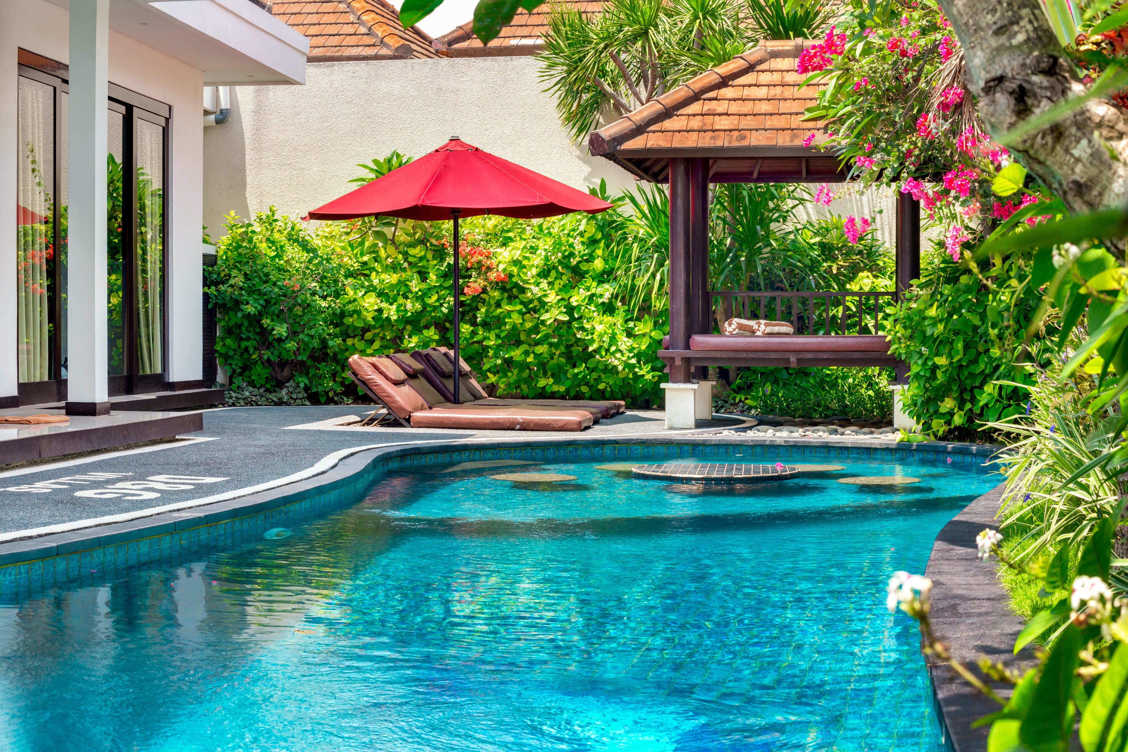 A swimming pool with lounge chairs and a red umbrella, surrounded by lush greenery and a tiled roof structure.