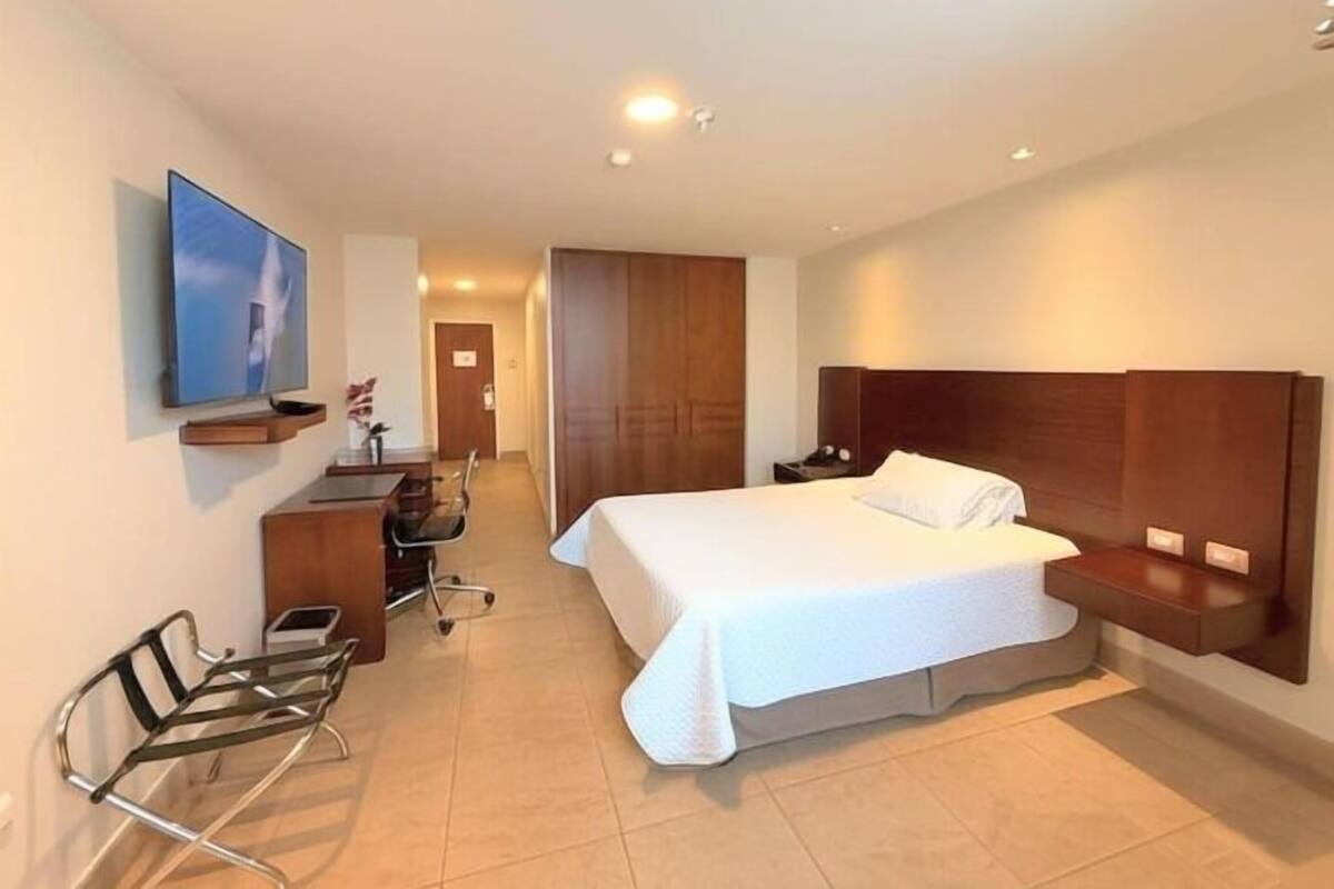 A modern hotel room with a large bed, a desk with a chair, a TV mounted on the wall, and a wooden wardrobe.