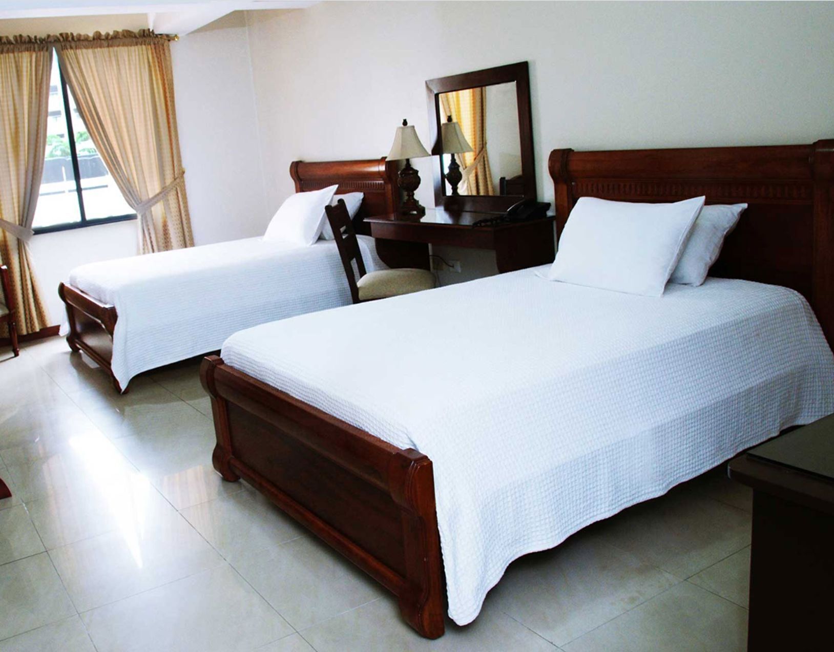 Dos camas de madera con ropa de cama blanca, una mesita de noche de madera, un escritorio con una lámpara y un espejo.