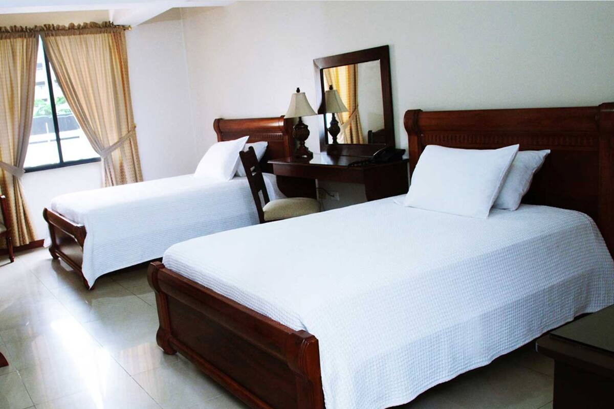 Two wooden beds with white bedding, a wooden nightstand, a desk with a lamp, and a mirror.